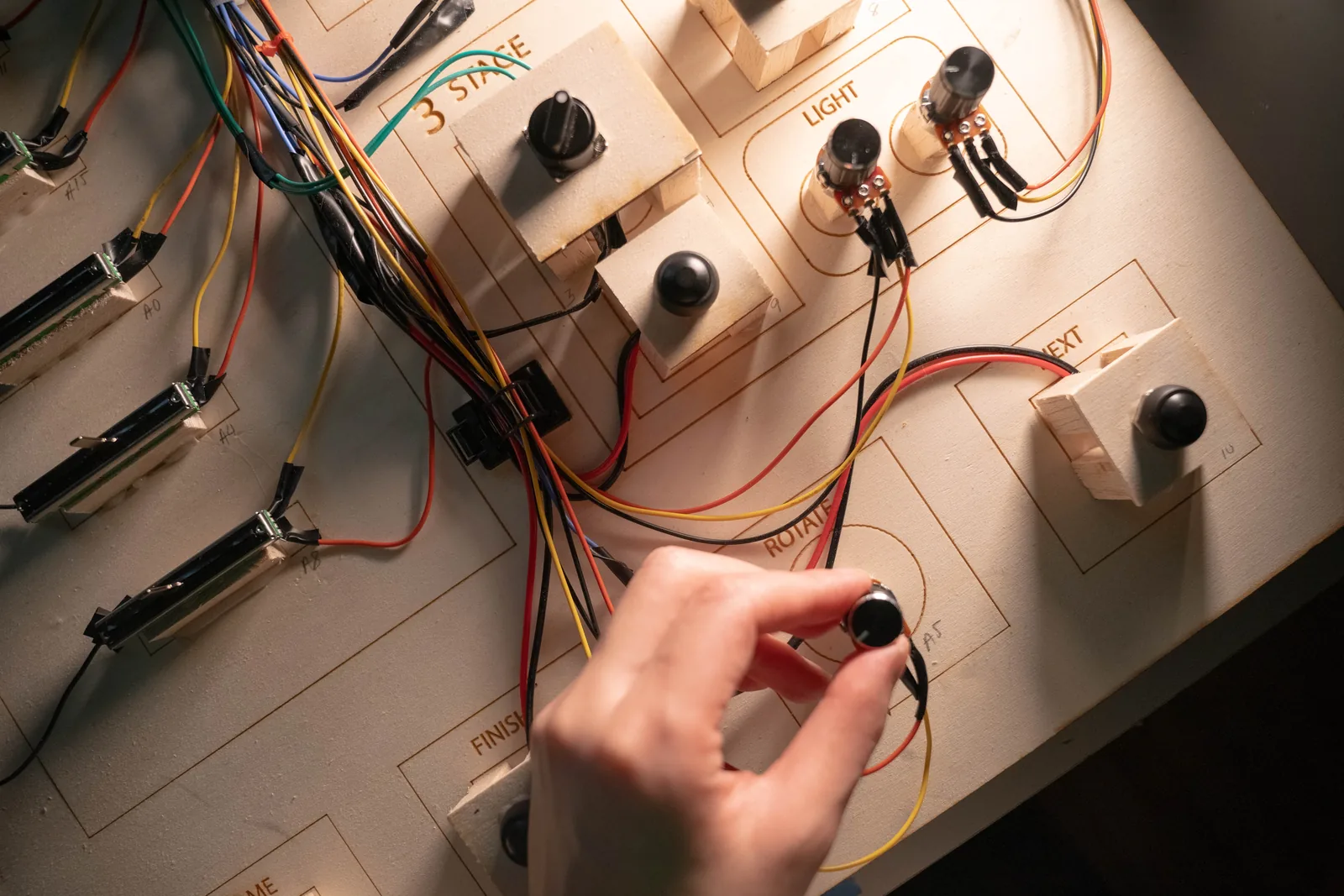 A hand on the prototype control panel, sliders and knobs in cardboard with wiring exposed and stage labels written by hand