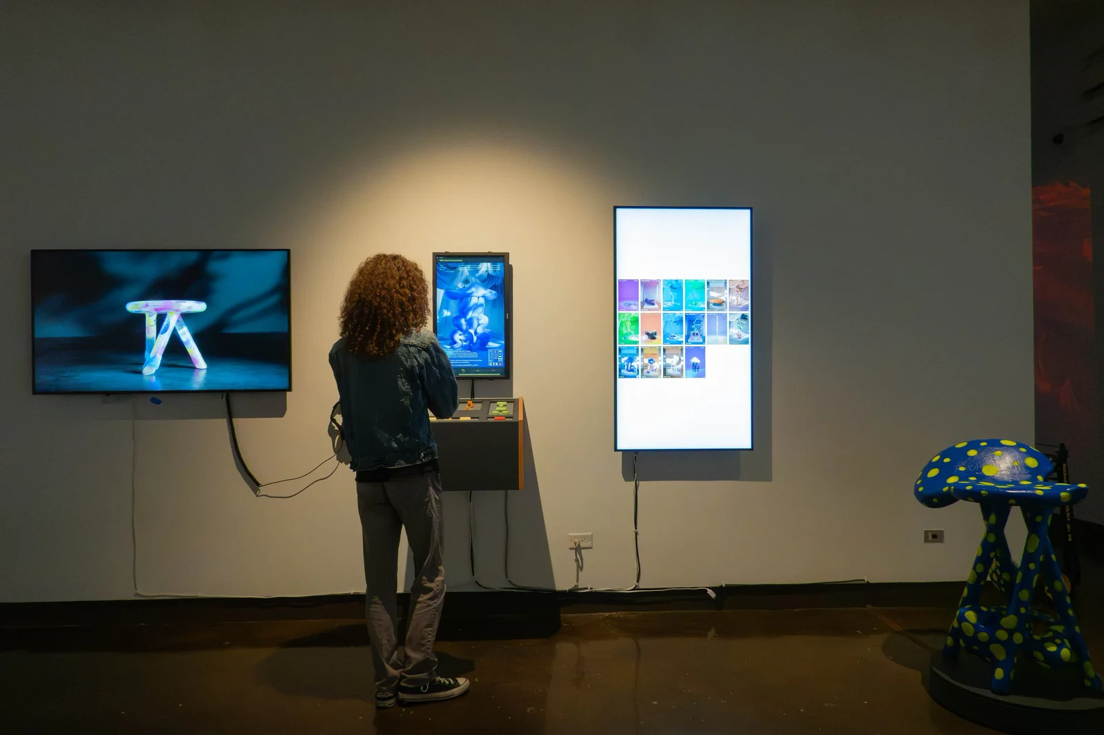 Installation view: a wall-mounted screen showing a rendered piece, the console with its interface screen, the gallery display, and the fabricated blue and yellow stool on a plinth