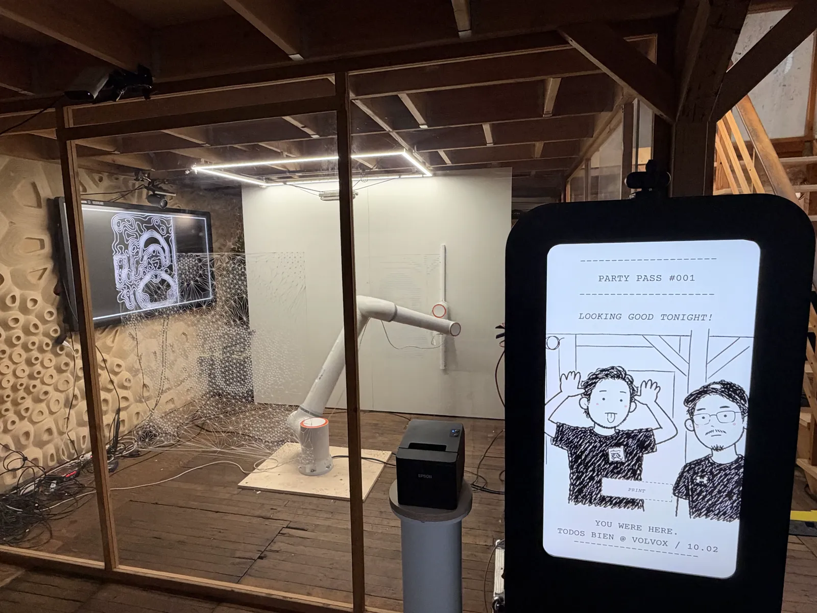 A photo booth screen showing a drawn portrait of two guests under the heading Party Pass #001, in front of a glass room with a white robot arm, a white wall and a TV of contour lines